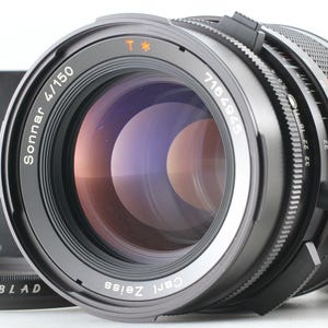 May include: A close-up of a Carl Zeiss Sonnar 4/150 camera lens. The lens is black with silver accents and has a large, reflective glass element. The lens is part of a Hasselblad camera system.