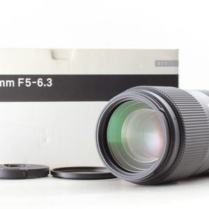 Puede incluir: Un objetivo Sigma 100-400mm F5-6.3 DG con un tamaño de filtro de 67 mm. El objetivo es negro con un anillo plateado y viene con una tapa de objetivo y una caja. La caja tiene el logotipo de Sigma e información del producto.
