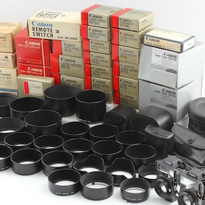 Pode incluir: Uma coleção de acessórios para câmeras Canon, incluindo parasóis, interruptores remotos e estojos para lentes. Os itens estão em suas embalagens originais, com caixas vermelhas, bege e brancas. Vários parasóis pretos e duas câmeras também são visíveis.