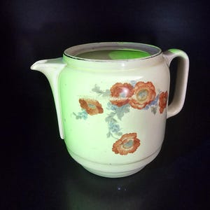 May include: Cream-coloured pitcher with a floral design featuring orange and red flowers with blue accents. The pitcher has a spout and a handle. The vintage-style pitcher is likely made of ceramic or porcelain.