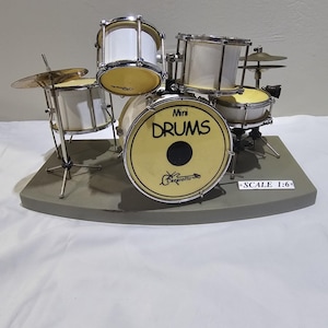 May include: Miniature drum set with white drums, yellow drum heads, and silver hardware. The bass drum features the word "DRUMS" in black. A cymbal and other drum components are also visible. The set is on a gray base, with "SCALE 1:6" printed on the base.