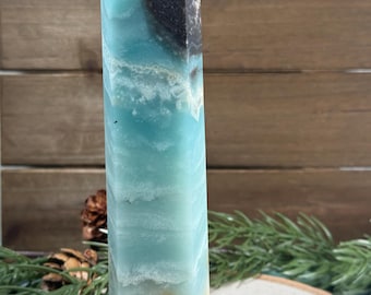 May include: A tall, polished crystal obelisk with a pointed top. The obelisk features a blend of light blue and white hues, with a dark brown patch near the top. The crystal is displayed against a wooden background with greenery.