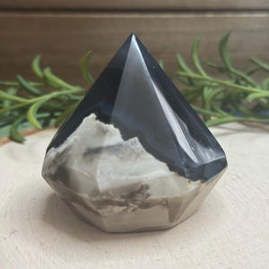 May include: A faceted, pyramid-shaped crystal with a polished, glossy finish. The crystal features a striking contrast of black and white bands, with a base of gray and tan hues. The gemstone is displayed on a wooden surface.