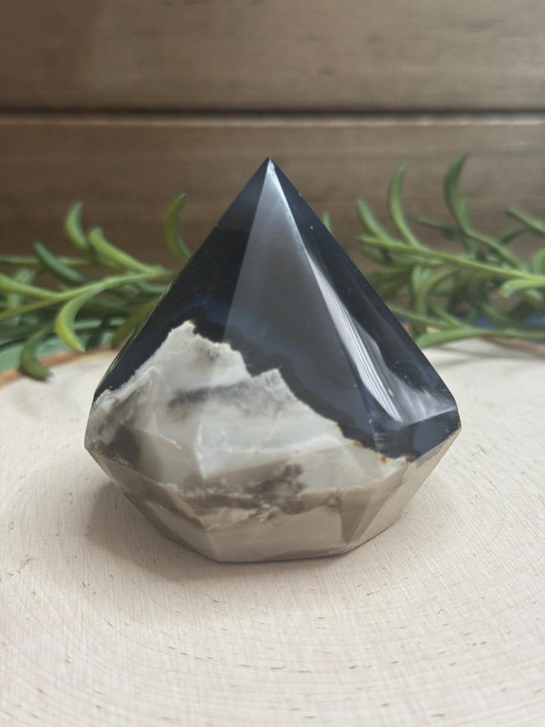 May include: A faceted, pyramid-shaped crystal with a polished, glossy finish. The crystal features a striking contrast of black and white bands, with a base of gray and tan hues. The gemstone is displayed on a wooden surface.