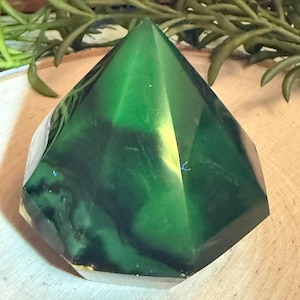 May include: A faceted, pyramid-shaped decorative object. The crystal is a deep green color with darker green marbling and a translucent quality. The base is a light, almost clear color. The object is displayed on a wooden surface.
