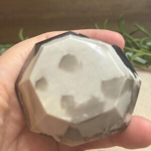 May include: A faceted, geometric stone with a mix of white, gray, and black tones. The stone is held in a hand, showcasing its polished surface and unique color variations. The shape is reminiscent of a geode or decorative object.