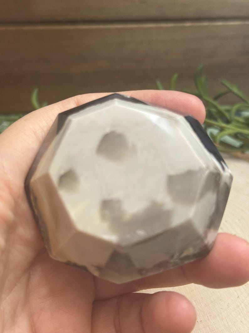 May include: A faceted, geometric stone with a mix of white, gray, and black tones. The stone is held in a hand, showcasing its polished surface and unique color variations. The shape is reminiscent of a geode or decorative object.