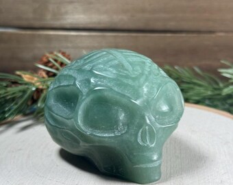 以下が含まれることがあります： 詳細な彫刻が施された緑色のクリスタルスカルの彫刻。 スカルは木の輪切りに展示されており、背景には常緑樹の小枝があります。 この彫刻は滑らかで磨かれた仕上がりで、ユニークな装飾品です。
