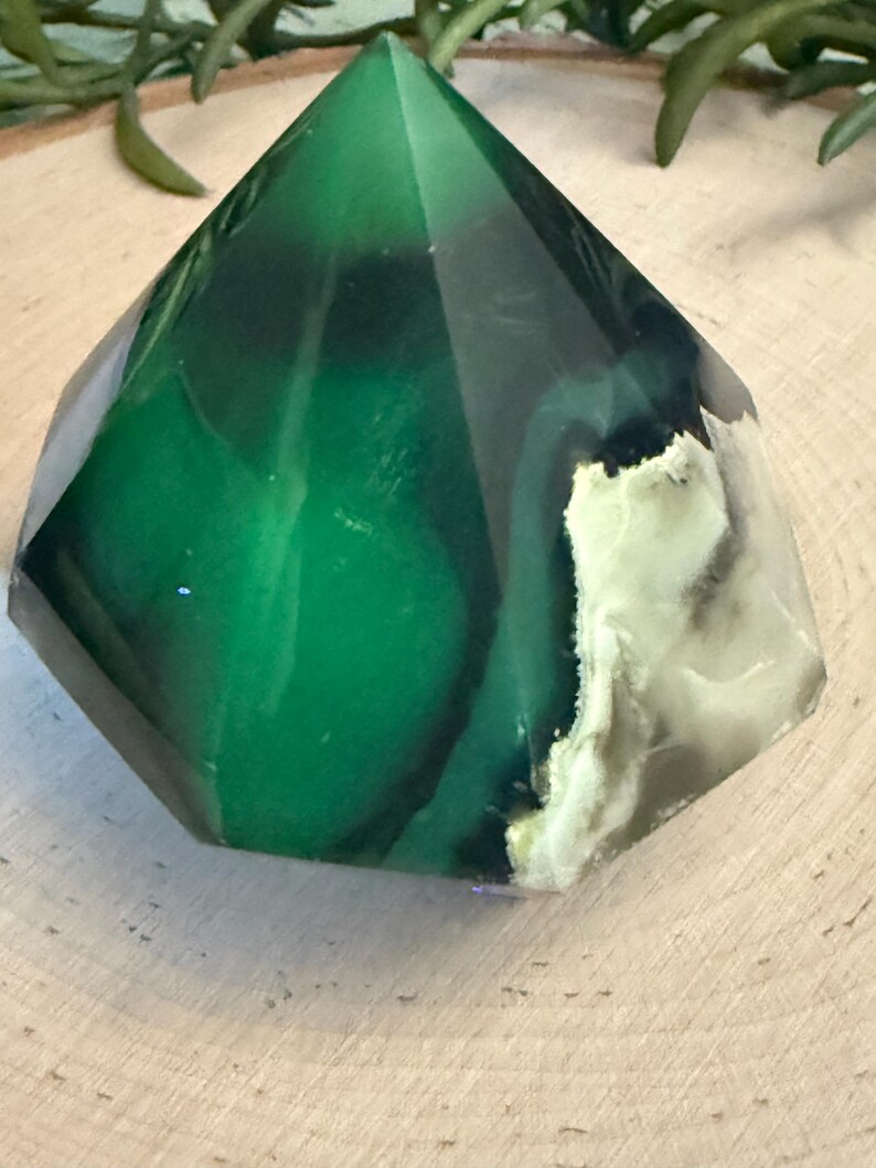 May include: A faceted, green crystal obelisk with a pointed top. The crystal has a mix of dark and light green hues, with some areas appearing translucent. The base has a white and gray pattern. The crystal is a decorative item.