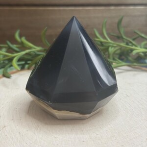 May include: A faceted, black obsidian crystal pyramid. The pyramid has a polished, reflective surface and a flat, light-colored base. The pyramid is displayed on a light-colored, textured wooden surface, with green sprigs in the background.