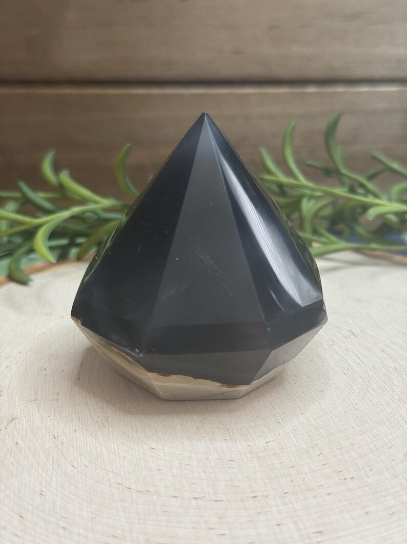 May include: A faceted, black obsidian crystal pyramid. The pyramid has a polished, reflective surface and a flat, light-colored base. The pyramid is displayed on a light-colored, textured wooden surface, with green sprigs in the background.