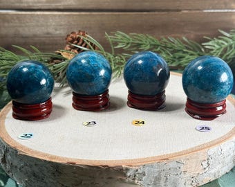 May include: Four blue-green crystal spheres on small, red wooden stands, displayed on a natural wood slice. Each sphere has a unique pattern. Numbers 22, 23, 24, and 25 are visible on the wood slice.