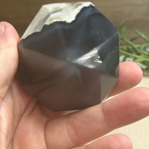 May include: A faceted, dark gray agate crystal with white and light gray banding at the top. The crystal is held in a hand, showcasing its polished surfaces and geometric shape. The crystal is a natural stone.