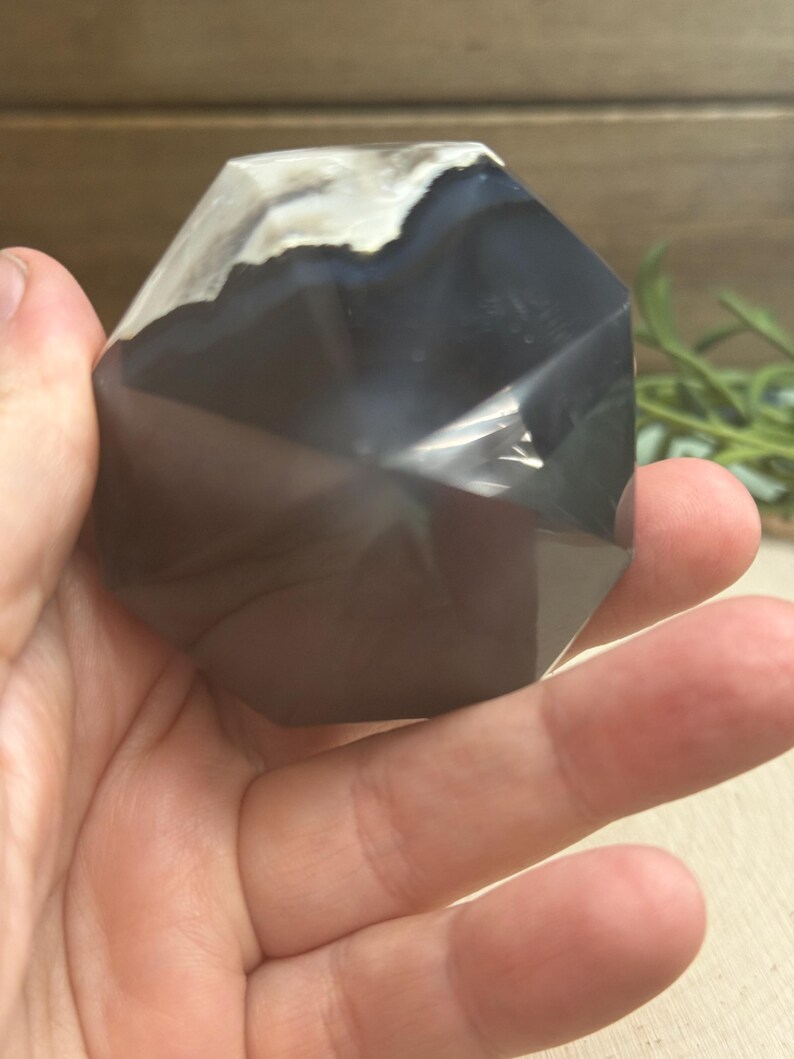May include: A faceted, dark gray agate crystal with white and light gray banding at the top. The crystal is held in a hand, showcasing its polished surfaces and geometric shape. The crystal is a natural stone.