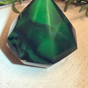 May include: A faceted, emerald-green crystal with a geometric shape. The crystal has a translucent quality, revealing darker green veining within. The base is a lighter, opaque color. The crystal sits on a light-colored wooden surface.
