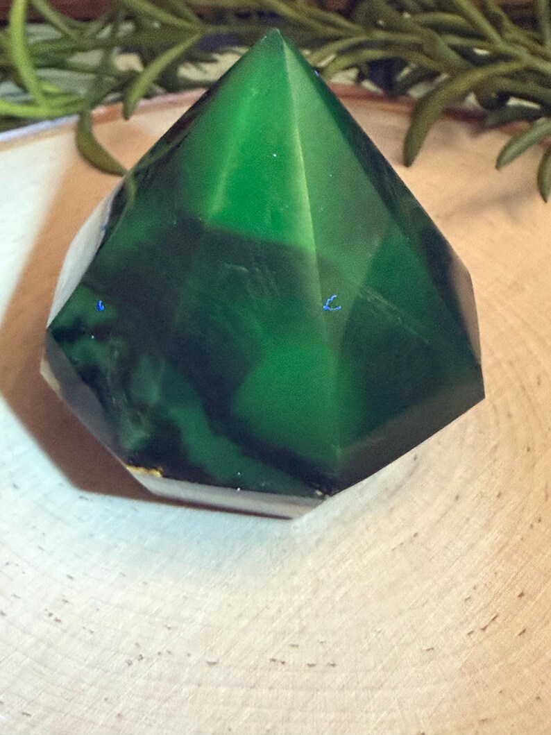 May include: A faceted, emerald-green crystal with a geometric shape. The crystal has a translucent quality, revealing darker green veining within. The base is a lighter, opaque color. The crystal sits on a light-colored wooden surface.