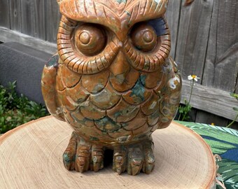 May include: A detailed, carved owl figurine in shades of brown, orange, and green. The owl is perched on a wooden slice, showcasing intricate feather and eye details. The sculpture is a decorative item, likely for home decor.