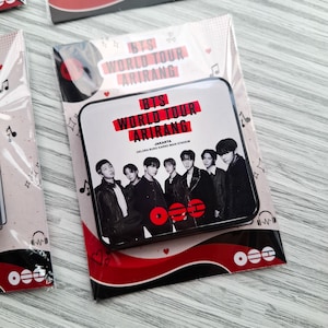 May include: A square metal pin featuring a black and white photograph of the seven members of BTS. The text reads BTS WORLD TOUR ARIRANG JAKARTA GELORA BUNG KARNO MAIN STADIUM. The design includes red accents, musical notes, and hearts.