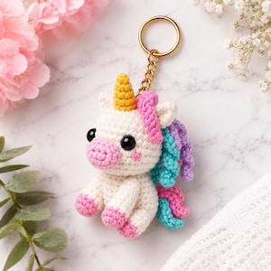 Könnte beinhalten: Ein handgefertigter gehäkelter Einhorn-Schlüsselanhänger. Das Einhorn ist weiß mit einem gelben Horn, einer rosa Schnauze und Hufen sowie einer bunten Mähne aus Rosa, Blau und Lila. Es hat schwarze Augen und einen goldfarbenen Schlüsselring.
