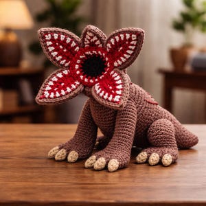 May include: A crocheted Demogorgon figure, featuring a detailed design with a red, white, and brown color scheme. The creature has a large, open flower-like head with sharp teeth and a dark center. The figure is posed on a wooden surface.