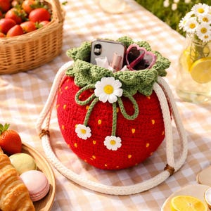 May include: A red, crocheted strawberry-shaped bag with a green leafy top and white daisy details. The bag has a white rope strap and contains a phone, sunglasses, and makeup. Strawberries and a picnic setting are in the background.