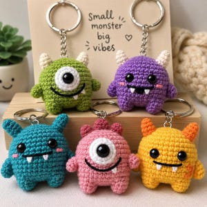 May include: A collection of five crocheted monster keychains in various colors: green, purple, blue, pink, and yellow. Each keychain features a unique design with horns, eyes, and a smiling mouth. The keychains are displayed with a sign that reads "Small monster big vibes."