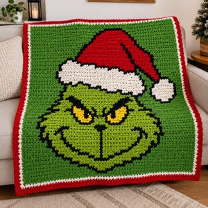 May include: A square crochet blanket featuring a green character with yellow eyes and a wide grin, wearing a red and white Santa hat. The blanket has a green background, a white inner border, and a thick red outer border with a textured finish.