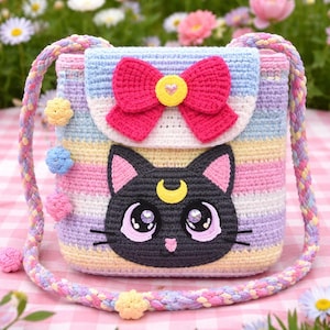 May include: A handmade crochet crossbody bag with a black cat face design. The bag features a pink bow, yellow heart, and a striped pattern in pastel colors. The strap and decorative elements are also crocheted.