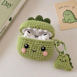 May include: A green crocheted dinosaur-shaped AirPod case with a matching keychain. The case has a smiling face with pink cheeks and black eyes. The AirPod case is open, revealing white AirPods.
