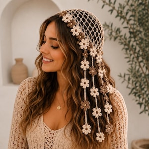 May include: A beige crochet head covering featuring a net pattern and a decorative border of small, alternating cream and brown flowers. Several long, dangling strands of matching crocheted flowers hang down from the side of the headpiece.