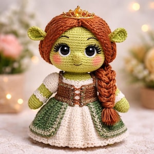 Op de afbeelding: Een handgemaakte gehaakte pop die lijkt op prinses Fiona uit de film Shrek. De pop heeft een groene huid, een rood gevlochten kapsel en een gouden kroon. Ze draagt een groene en witte jurk met een bruin lijfje. De pop is ongeveer 20 cm hoog.