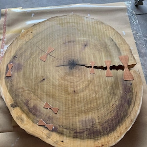Puede incluir: Una losa de madera grande y redonda con anillos de crecimiento visibles y un borde natural. La losa tiene varias grietas rellenas con pequeñas incrustaciones decorativas en forma de mariposa hechas de madera de color más claro. La madera tiene un color cálido y natural.