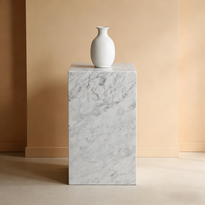 以下が含まれることがあります： 白いセラミックの花瓶が、大理石の台座の上に置かれています。花瓶は丸みを帯びたボディと細い首を持っています。台座は灰色の縞模様が入った正方形のブロックです。背景はニュートラルなベージュ色です。