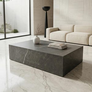Puede incluir: Una mesa de centro rectangular de mármol gris oscuro con un patrón de vetas blancas. Un pequeño jarrón blanco con una ramita y dos libros están sobre la mesa. La mesa está en un salón moderno.