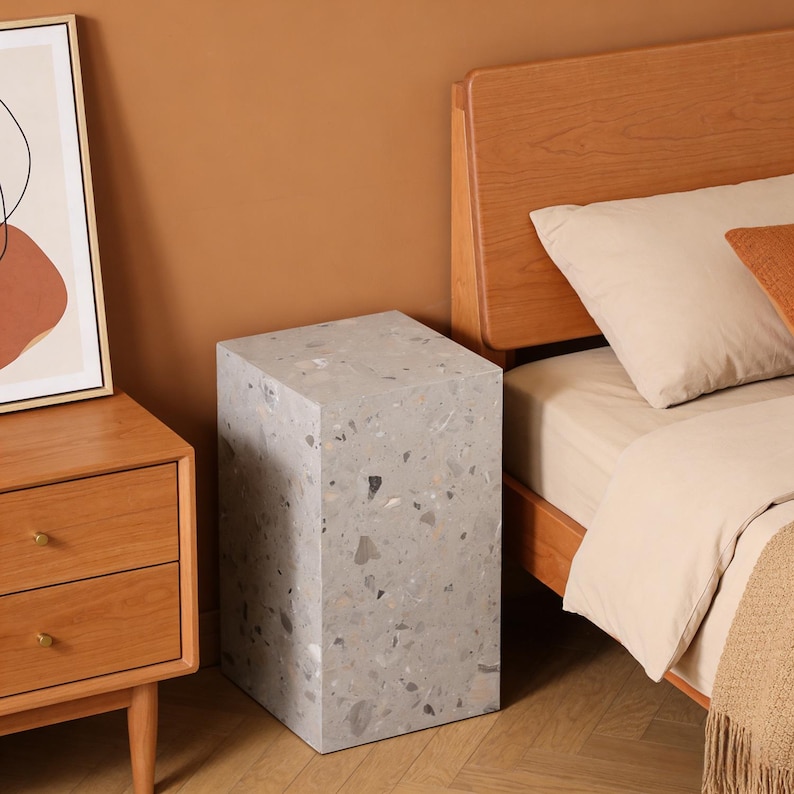 以下が含まれることがあります： テラゾーデザインのグレーの長方形サイドテーブル。テーブルは無垢材でできており、表面は滑らかです。テーブルは木製のナイトスタンドとベージュの掛け布団が付いたベッドの隣にあります。