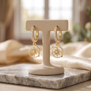 Puede incluir: Un par de pendientes de aro dorados con diseño de brújula. Los aros están incrustados con pequeñas piedras transparentes. Cada pendiente presenta un colgante circular con una rosa de los vientos y una sola piedra pequeña y transparente. Los pendientes se exhiben en un soporte beige.