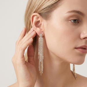 Pode incluir: Brincos compridos dourados com borlas e cristais transparentes brilhantes. Os brincos apresentam uma parte superior retangular com vários fios de cristais em cascata, criando um visual glamouroso e elegante. Os brincos são projetados para balançar e captar a luz.