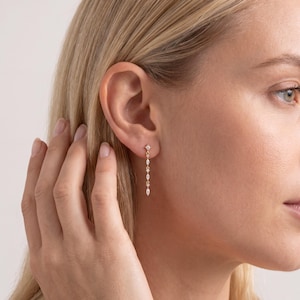 May include: A pair of gold earrings with a delicate design. Each earring features a small square stud at the top, with a series of small, sparkling, teardrop-shaped gems suspended below. The earrings are worn in the ear of a person with blonde hair.