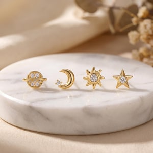 May include: Four gold-colored stud earrings with celestial designs. The set includes a planet, crescent moon, sun, and star, each adorned with small, clear gemstones. The earrings are displayed on a white marble surface.
