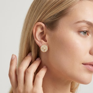 Puede incluir: Pendientes de botón dorados con diseño de rosa de los vientos. Los pendientes redondos tienen una estrella central y un borde de pequeñas piedras brillantes. Los pendientes se muestran en la oreja y la cara de una persona.