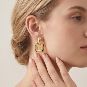 May include: Gold-coloured, square earrings with a rectangular top detail. The earrings have a polished, reflective surface. The image shows the earrings worn on a person's ear, with the focus on the jewelry.