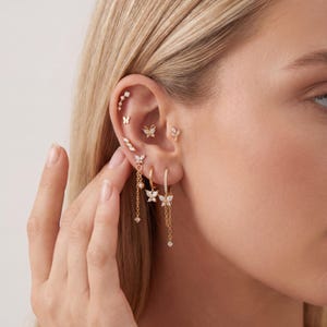 May include: A collection of gold-toned earrings and ear piercings, including butterfly and geometric designs. The earrings feature small, sparkling gemstones. The jewelry is displayed on a person's ear, showcasing the various styles and placements.