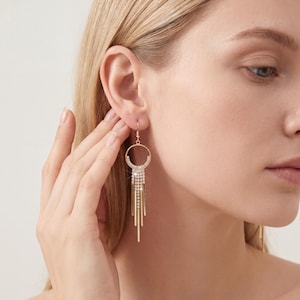 Puede incluir: Pendientes colgantes dorados con una parte superior circular y acentos lineales en cascada. Los pendientes presentan un diseño brillante con cristales en la parte superior y barras doradas colgantes. Los pendientes están diseñados para orejas perforadas.