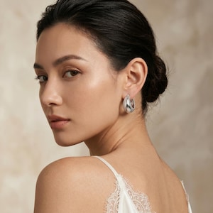 May include: A woman wearing silver hoop earrings. The earrings have a smooth, polished finish with a row of small, sparkling stones along the inner edge. The earrings are a teardrop shape and reflect the light.
