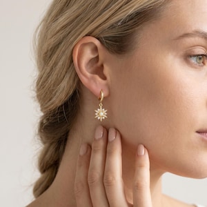 May include: A gold-colored earring with a starburst design, featuring a central white stone and surrounding clear stones. The earring is attached to a small hoop. The image shows the earring on a person's ear.
