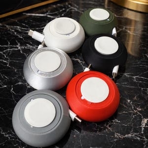 May include: A collection of colorful, round cable organizers in white, silver, gray, red, black, and olive green. Each organizer has a white center and a retractable cable with a USB-C connector. The organizers are on a black marble surface.