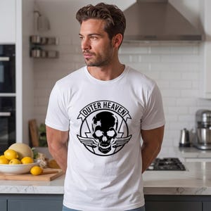 Könnte beinhalten: Ein weißes kurzärmeliges T-Shirt mit einem schwarzen Grafikdruck auf der Brust. Das Design zeigt einen zentralen Totenkopf mit Flügeln, der von einem kreisförmigen Emblem mit dem Text OUTER HEAVEN in fetter Schrift umgeben ist.