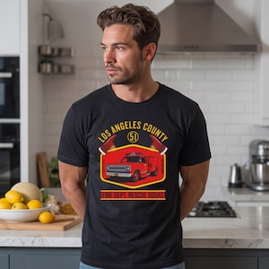 May include: A black short-sleeved t-shirt featuring a graphic of a red fire rescue truck inside a yellow and red emblem. The text reads LOS ANGELES COUNTY 51 FIRE DEPT. RESCUE SQUAD. The design includes two red fire helmets above the truck.