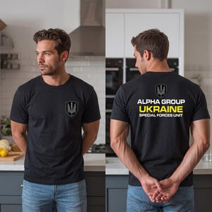 May include: A black short-sleeve t-shirt featuring a small emblem on the left chest and a large graphic on the back. The back design displays a shield emblem above the text ALPHA GROUP UKRAINE SPECIAL FORCES UNIT in white and yellow lettering.