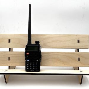 May include: A black handheld two-way radio with an antenna stands on a small wooden bench. The bench is made of light-colored wood and has a simple design with visible joinery. The radio has a display screen and a keypad.